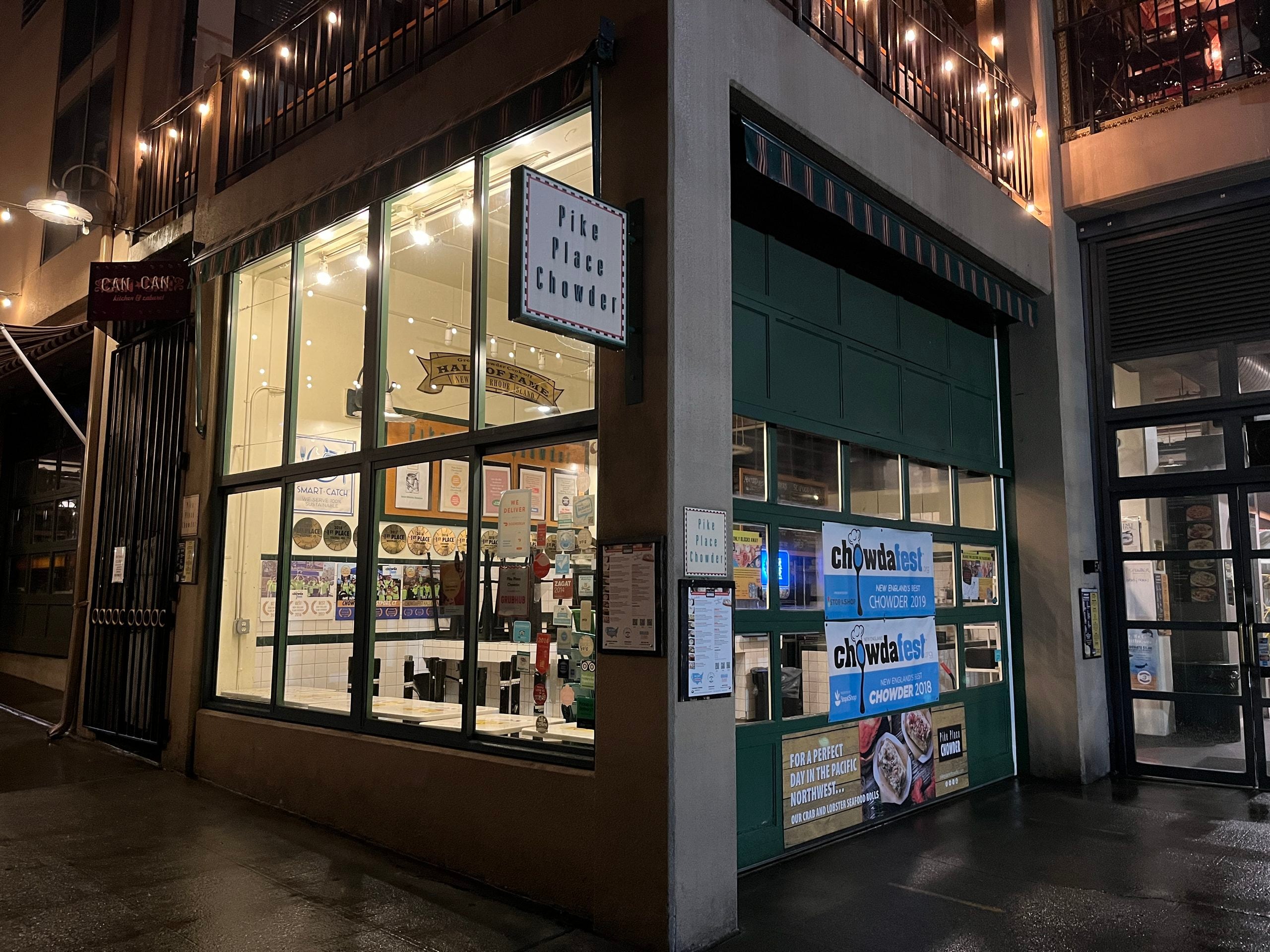 Exterior of the Pike Place Chowder restaurant at night, showing large windows, signage, and awards posted on the glass.