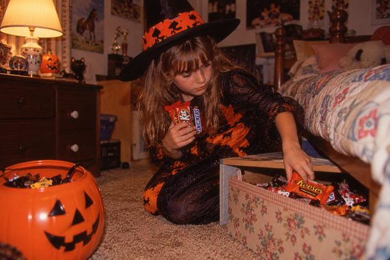 A young girl in a witch costume kneels on the floor, holding a KitKat and Snickers bar while reaching into a bin filled with candy.