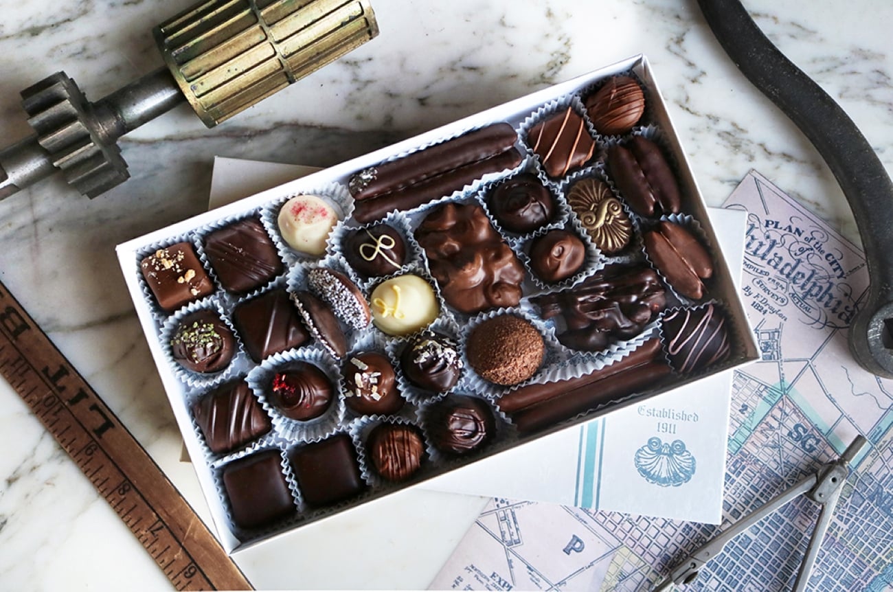 A rectangular box of assorted chocolates, the best souvenir, surrounded by vintage drafting tools and a map of Philadelphia.