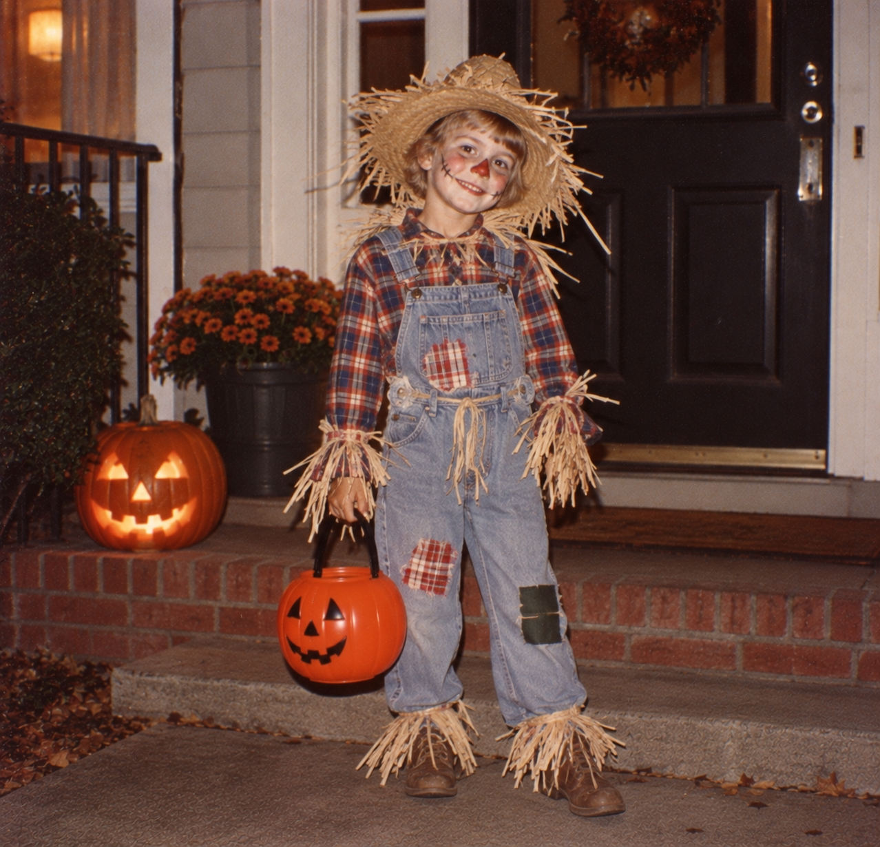 A child dressed as a scarecrow in plaid, overalls, and a straw hat stands on a porch holding a plastic jack-o’-lantern pumpkin bucket.
