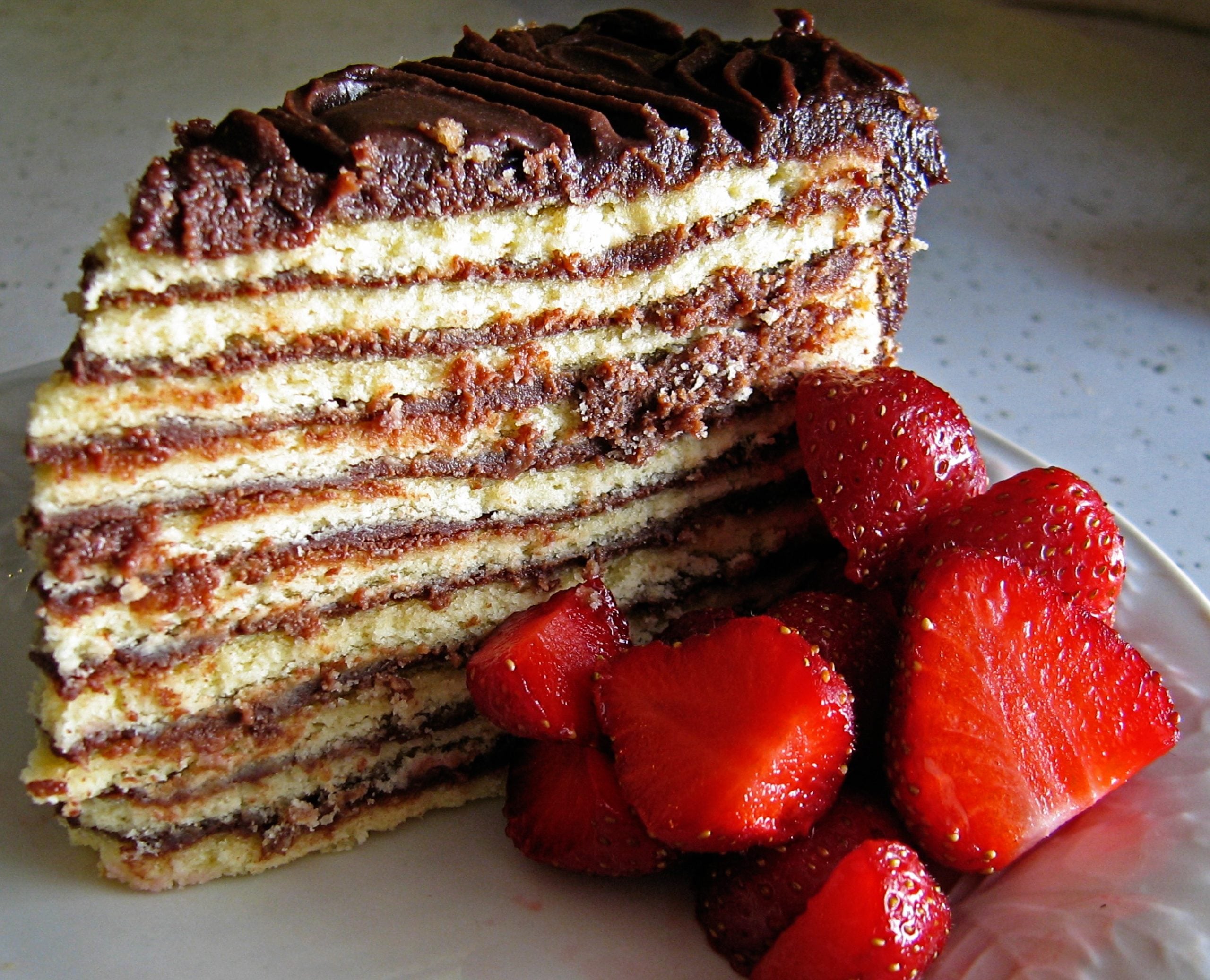 A slice of multi-layered cake with chocolate filling and frosting, served with fresh strawberries on a white plate.
