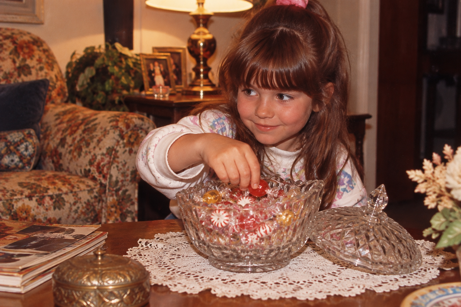 A young girl reaches into a cut-glass candy bowl on a lace-covered table to pick up a red piece of candy.