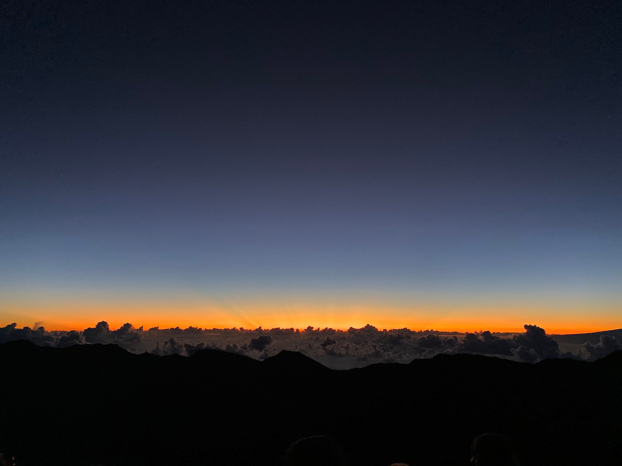 A sunrise glowing above a dark mountain range and a layer of clouds.