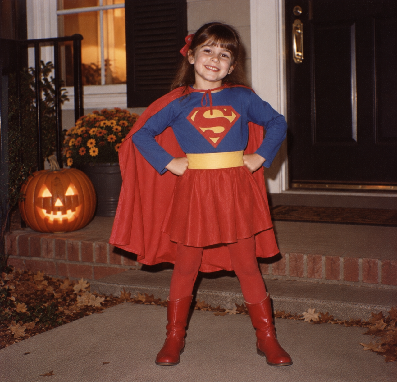 A young girl dressed in a Supergirl costume standing on a porch with a lit jack-o’-lantern.