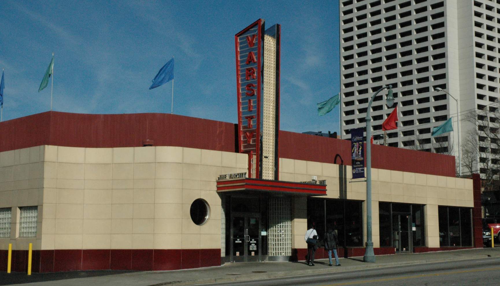 The Varsity restaurant with its iconic red vertical sign, exterior walls in red and cream, and a tall building in the background.