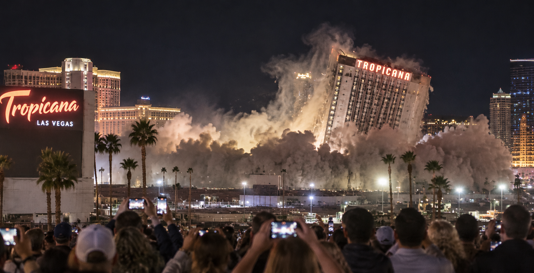A crowd watches and records as the Tropicana hotel in Las Vegas is demolished at night, with dust and debris rising—an echo of Old Las Vegas Traditions—amid city lights and palm trees.