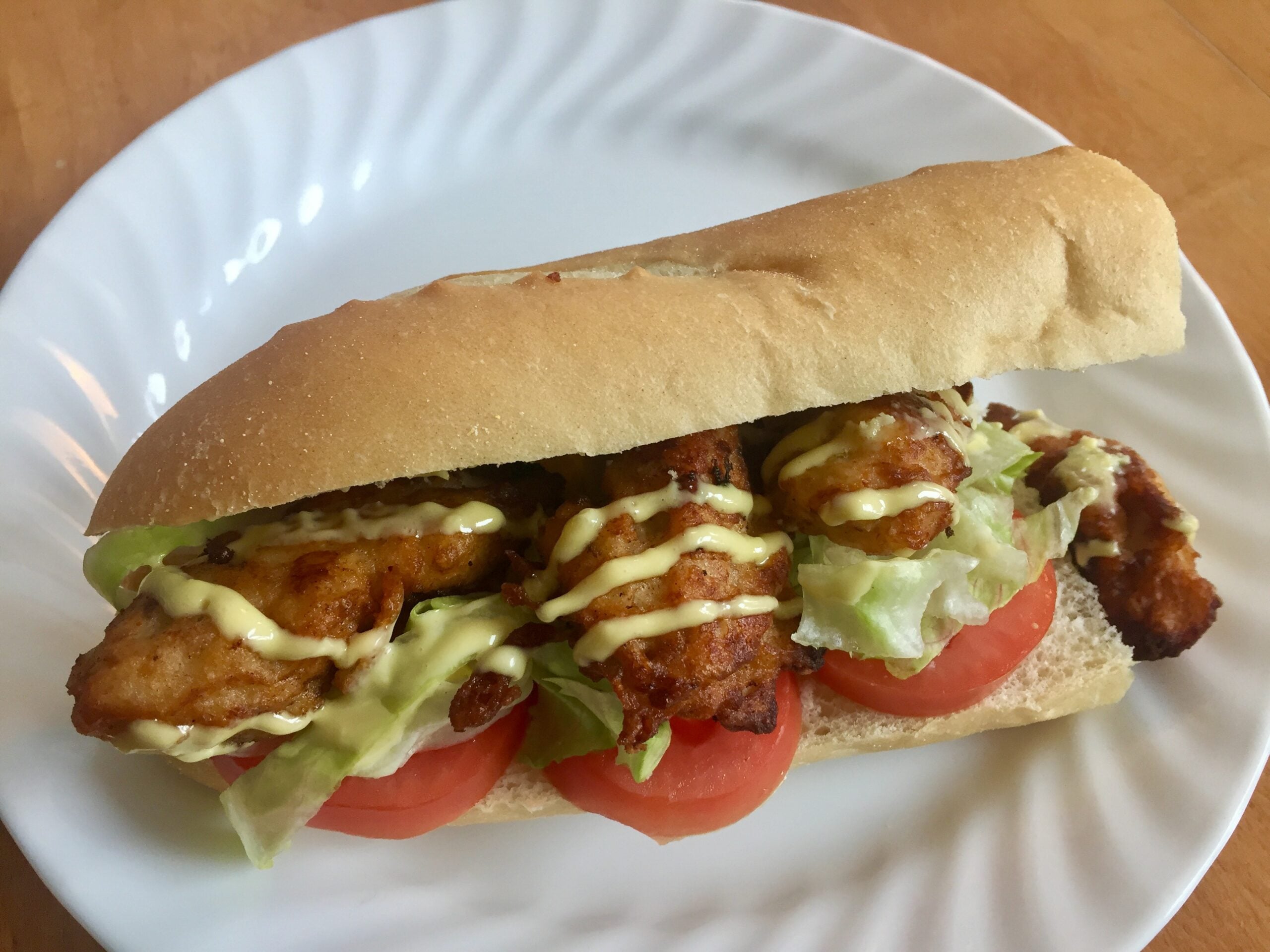 A fried chicken sandwich on a white plate with lettuce, tomato, and a drizzle of yellow sauce.