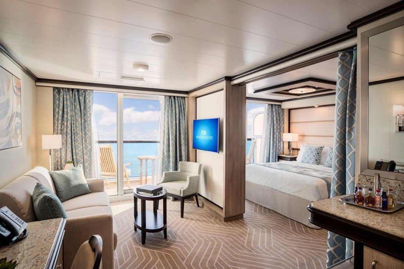 Cruise ship suite featuring a living area with a sofa, a bedroom with a bed, and a private balcony overlooking the ocean.