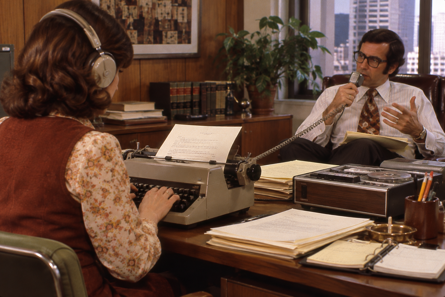 A woman with headphones types on a typewriter while a man at a desk speaks into a handheld microphone.
