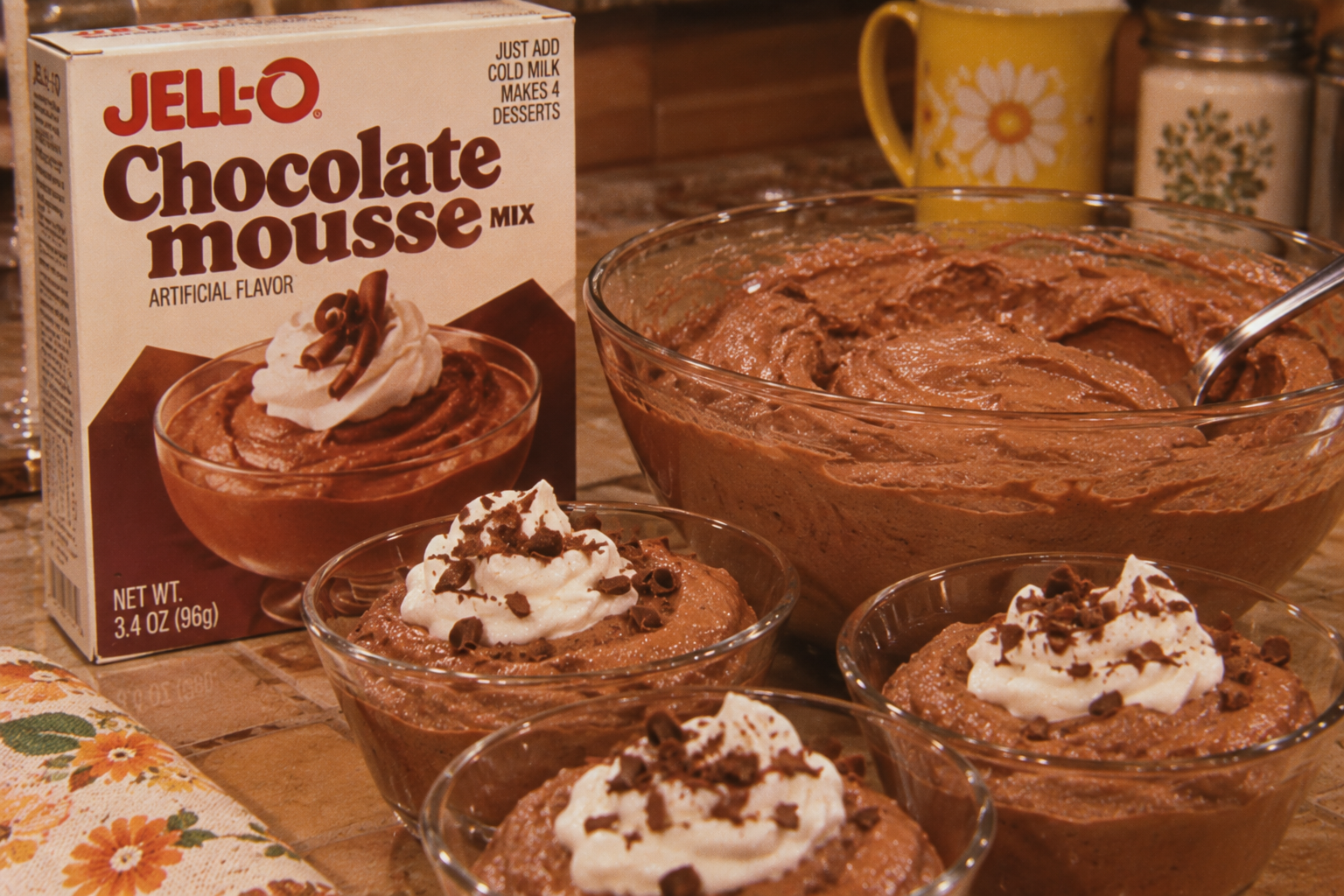 A box of Jell-O chocolate mousse mix sits next to a large bowl and three small bowls filled with whipped chocolate mousse, topped with whipped cream and chocolate shavings.