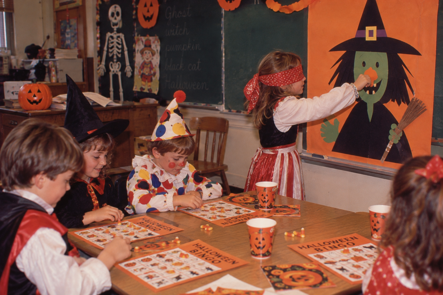 A child wearing a blindfold pins a nose onto a paper witch on a classroom wall while other children in costumes play Halloween bingo at a table.