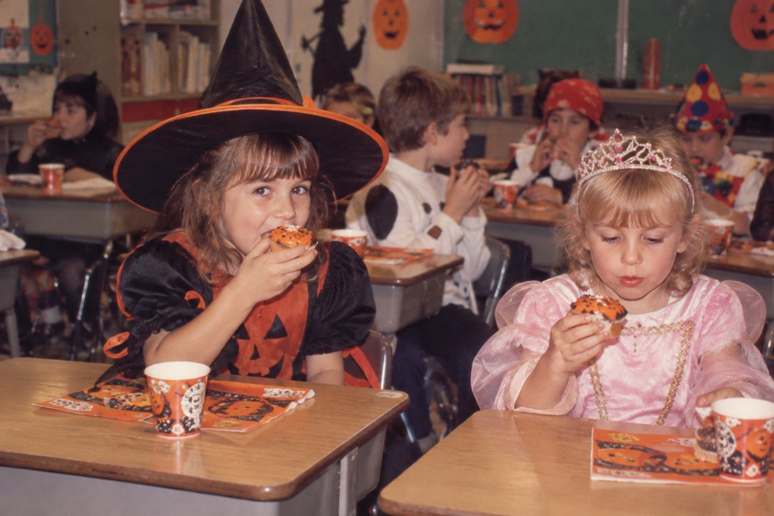Two children in Halloween costumes eat cupcakes at their classroom desks. Halloween at School in the '80s.