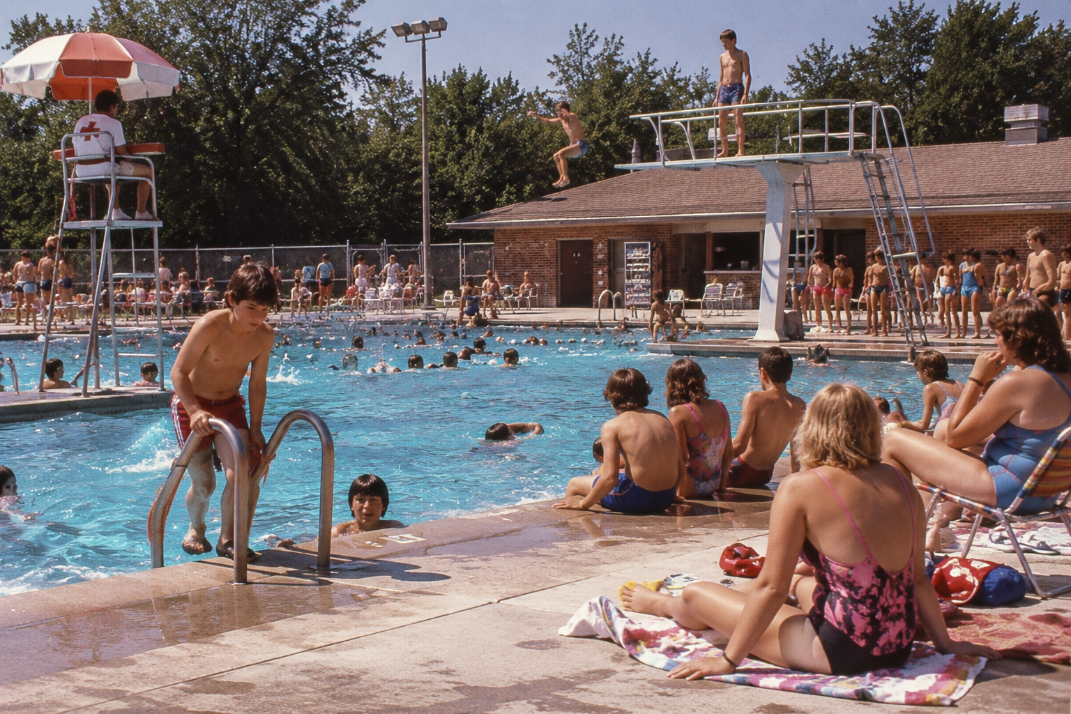 Many people swim and sunbathe at a crowded outdoor public pool, where one person jumps from a high diving board.