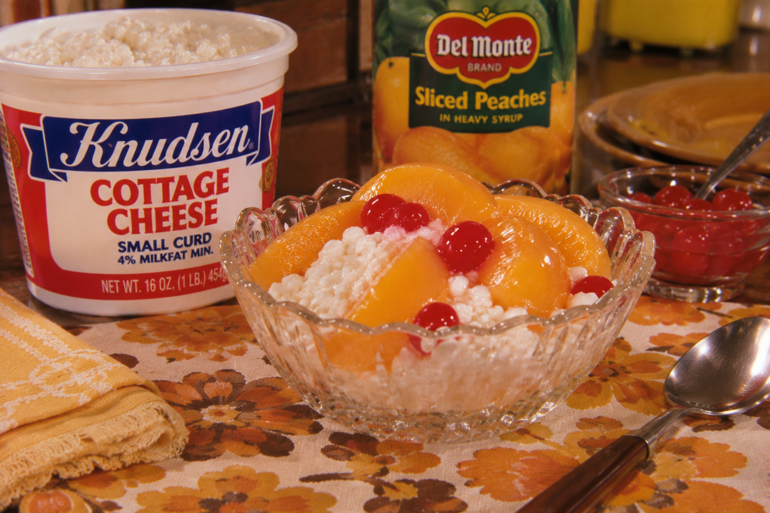 A glass bowl with cottage cheese, peach slices, and cherries sits on a floral tablecloth. Nearby are a spoon, a cottage cheese container, a can of peaches, a napkin, and a small bowl of cherries.