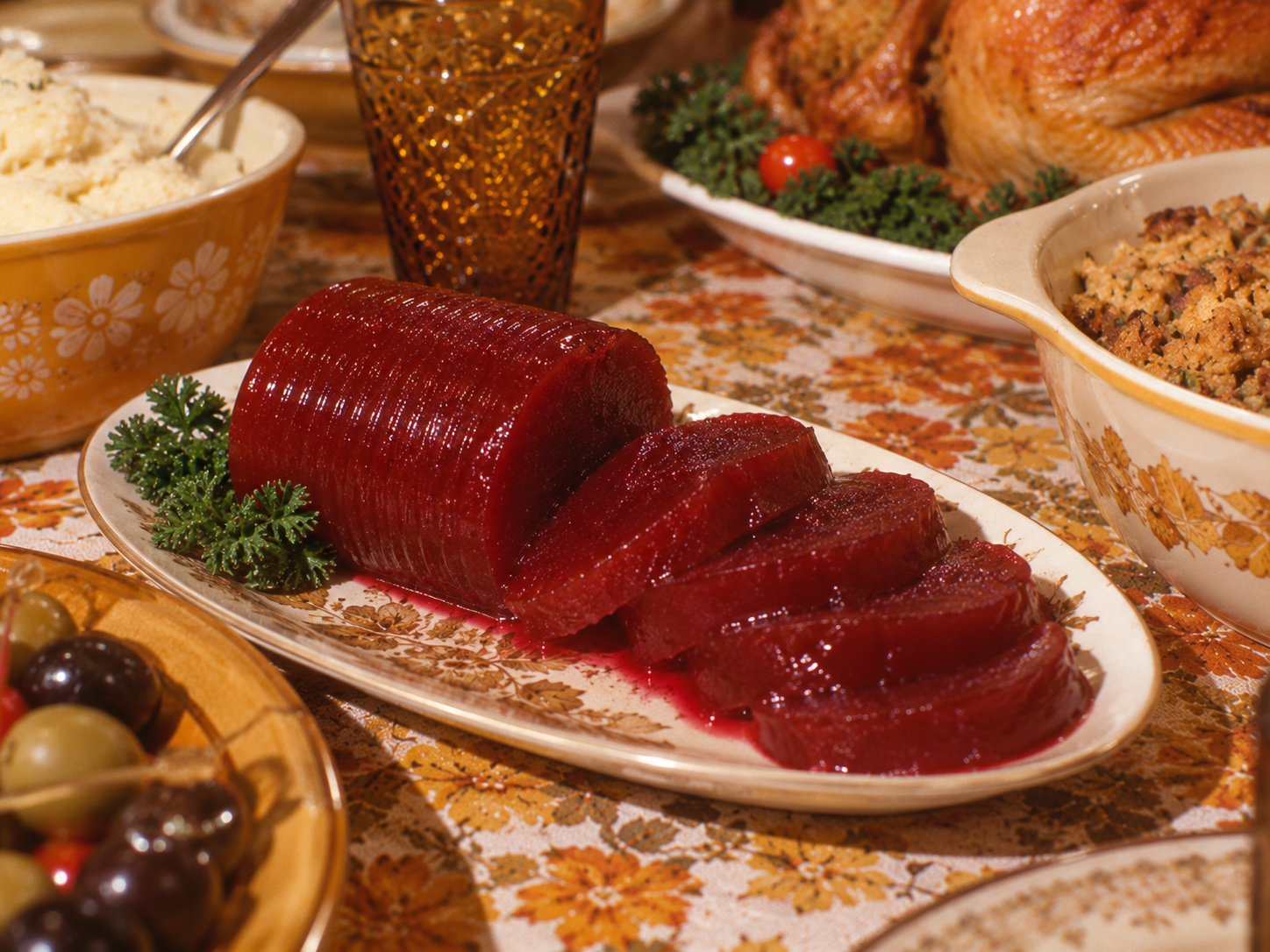 A cylinder of jellied cranberry sauce with several slices cut off, served on a floral platter surrounded by other holiday dishes.