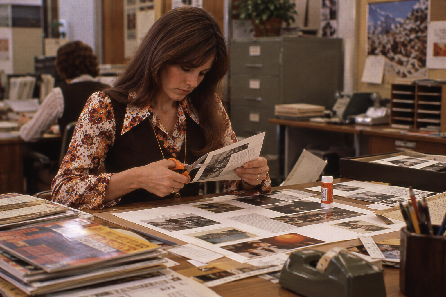 A woman in a floral blouse and dark vest cuts a photograph with orange-handled scissors at an office desk covered with documents and photo layouts.