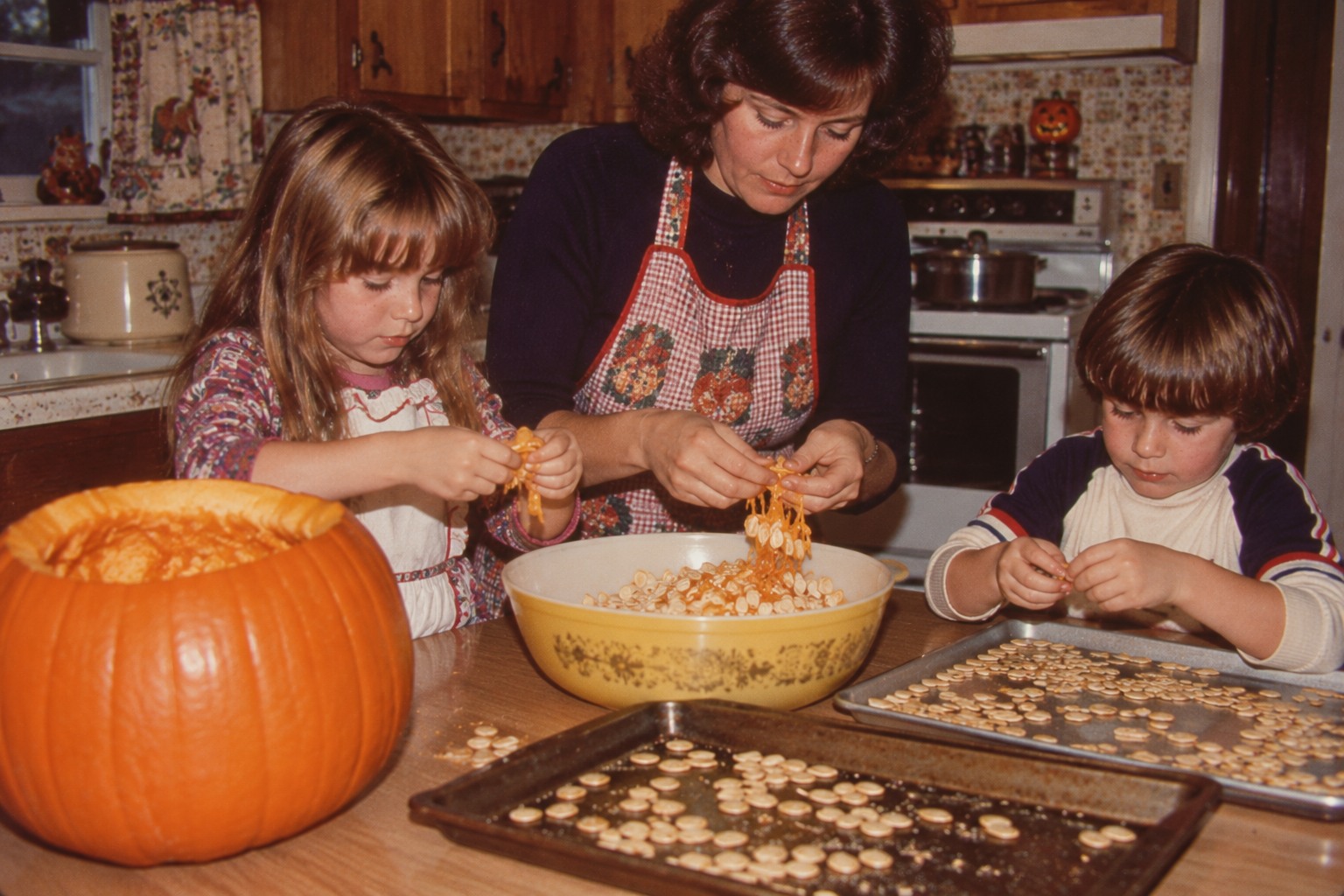 A woman and two children sitting at a kitchen table cleaning pumpkin seeds into a bowl, with trays of prepared seeds nearby.