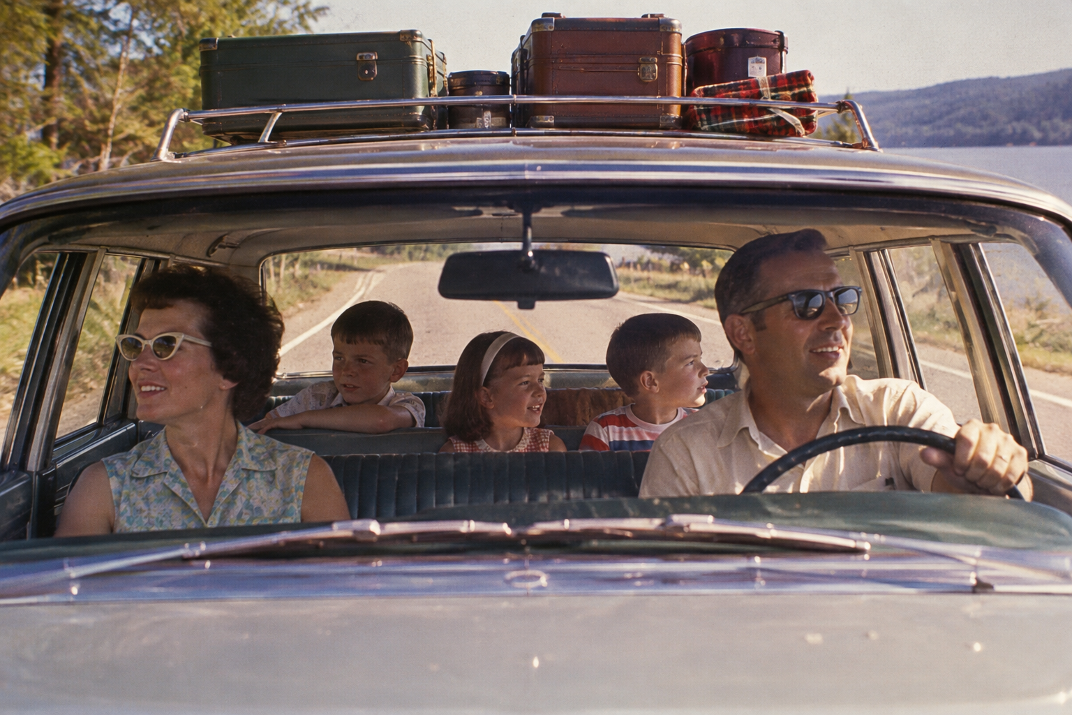A family of four riding in a car on a road trip, with suitcases and a blanket strapped to the roof rack.