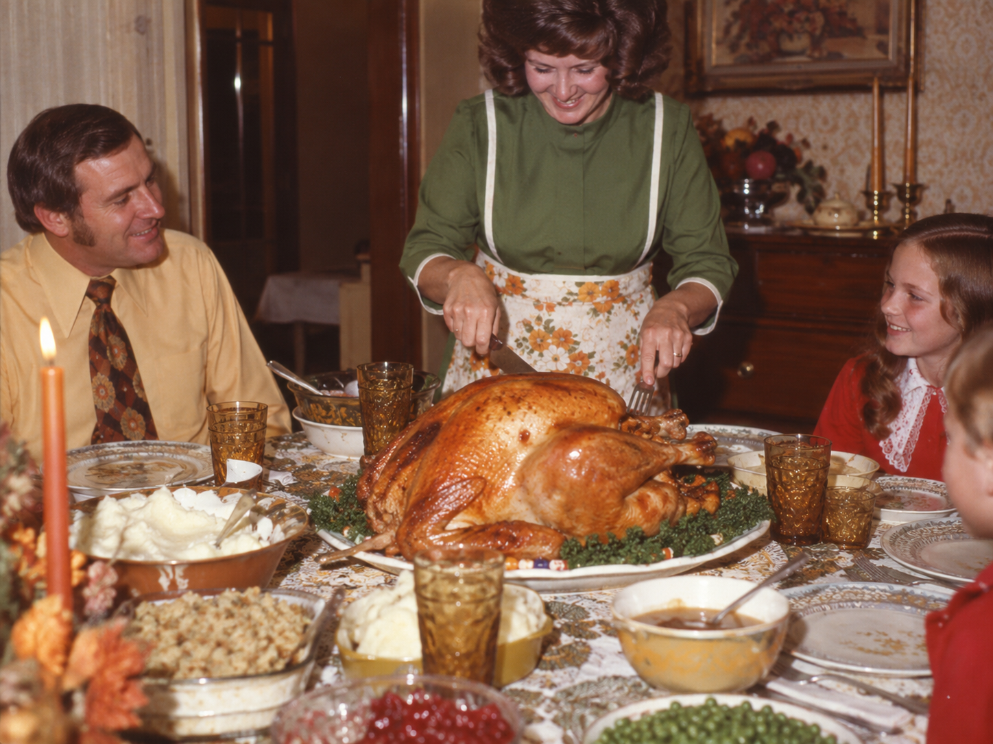 A woman in a green apron carves a large roasted turkey at a dinner table while a man and a girl look on.