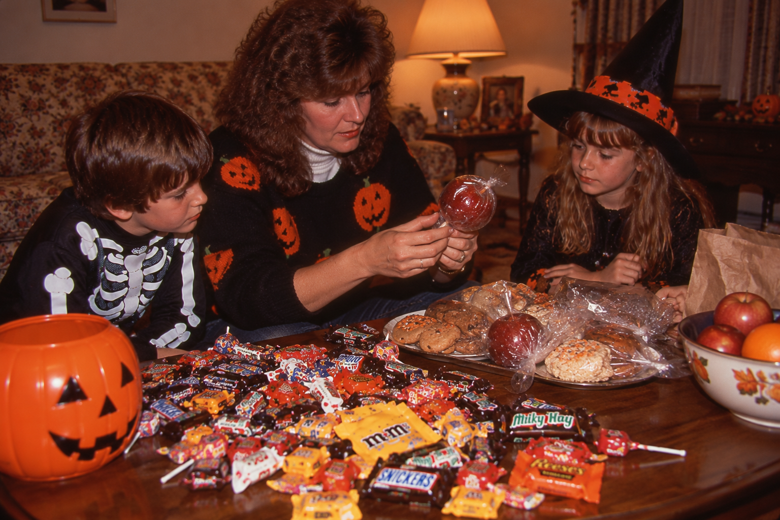 A woman and two children in Halloween costumes sit at a table covered in a large pile of candy and wrapped treats.