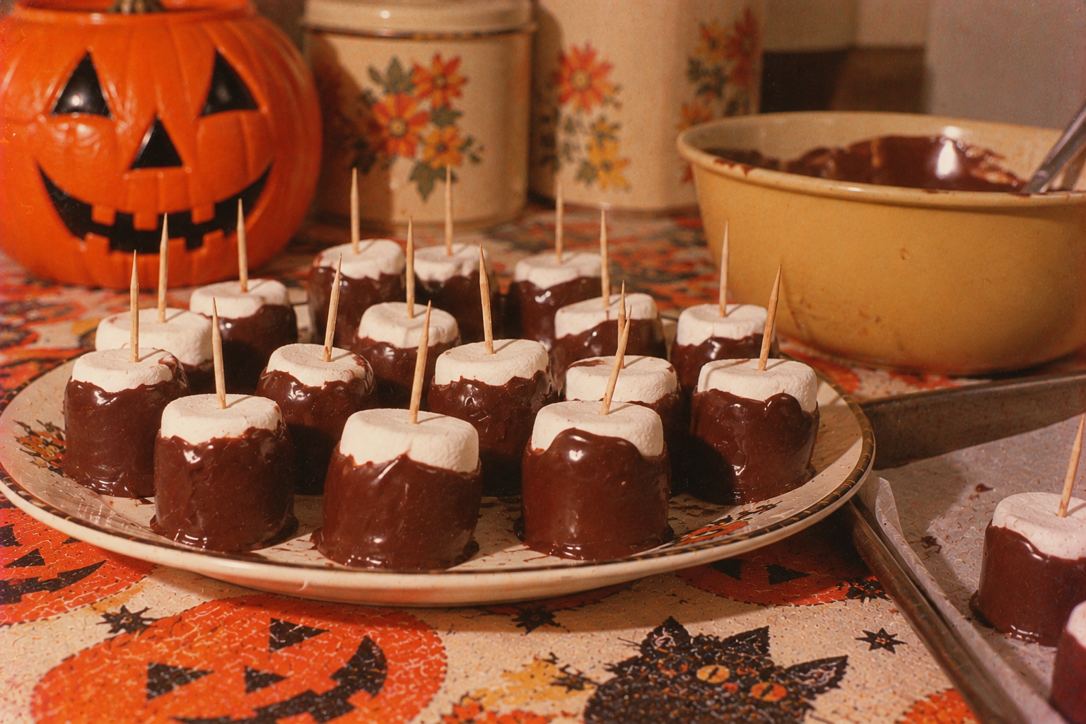 A plate of chocolate-dipped marshmallows, each with a toothpick inserted, sitting on a Halloween-themed tablecloth next to a jack-o’-lantern and a bowl of chocolate.