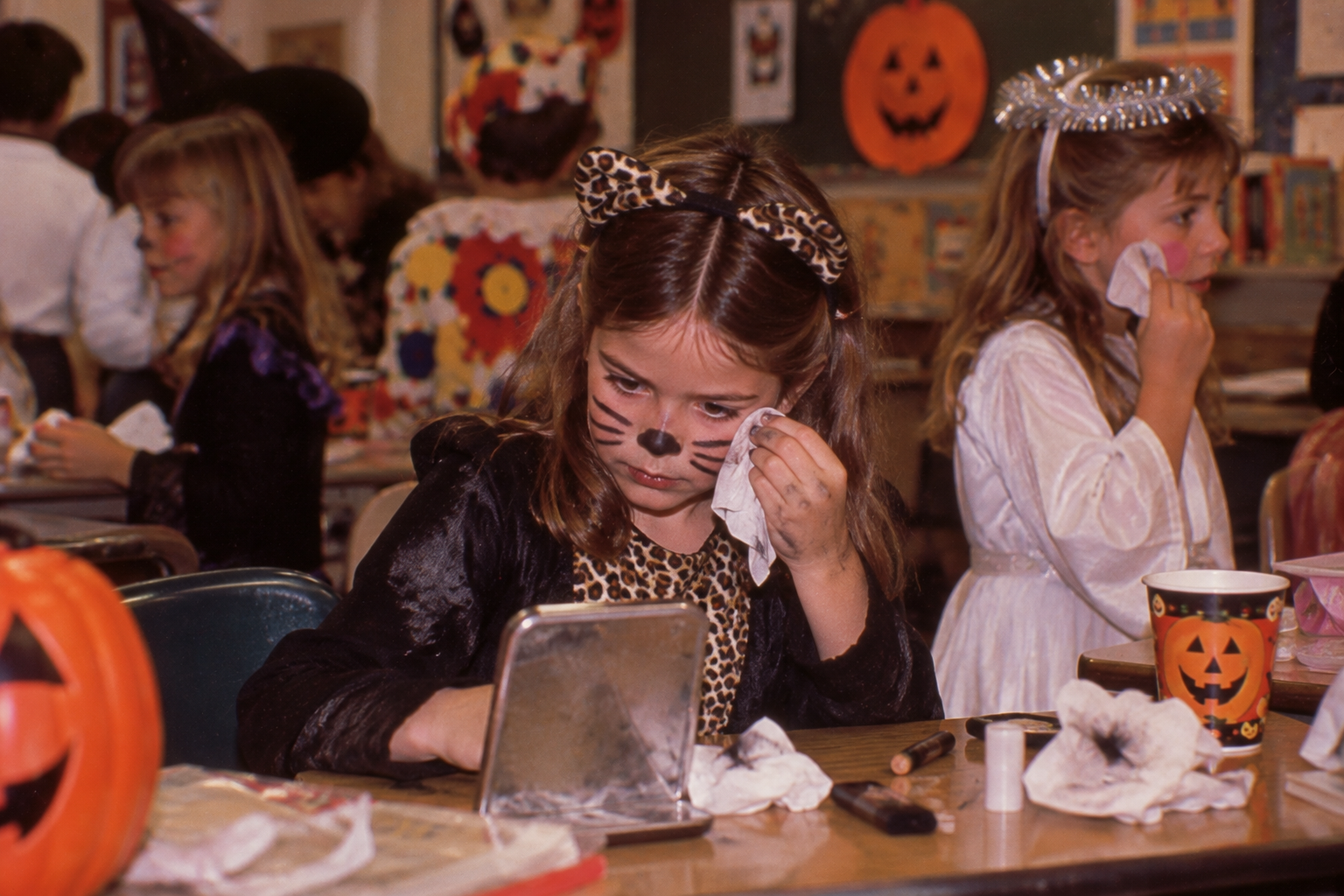 A young girl in a leopard costume and cat face paint looks into a small mirror while wiping her face with a tissue at a classroom Halloween party.