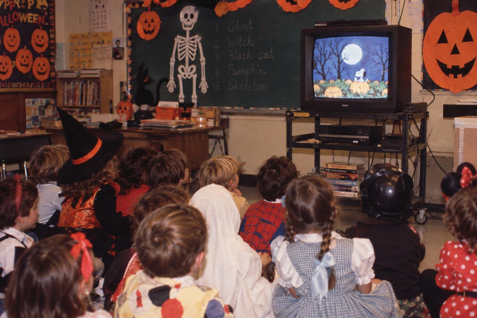 A group of children in Halloween costumes sit on a classroom floor watching a “Peanuts” holiday special on a television screen.