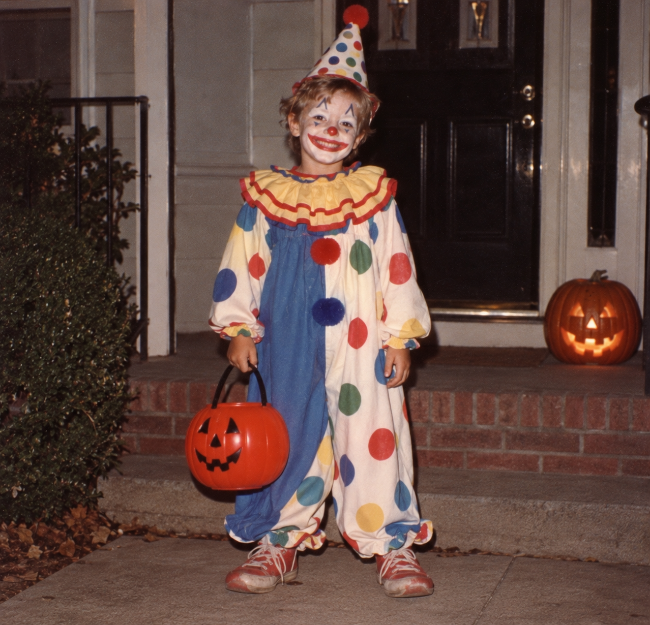 A child in a clown costume and makeup standing on a porch with a plastic pumpkin trick-or-treat bucket.