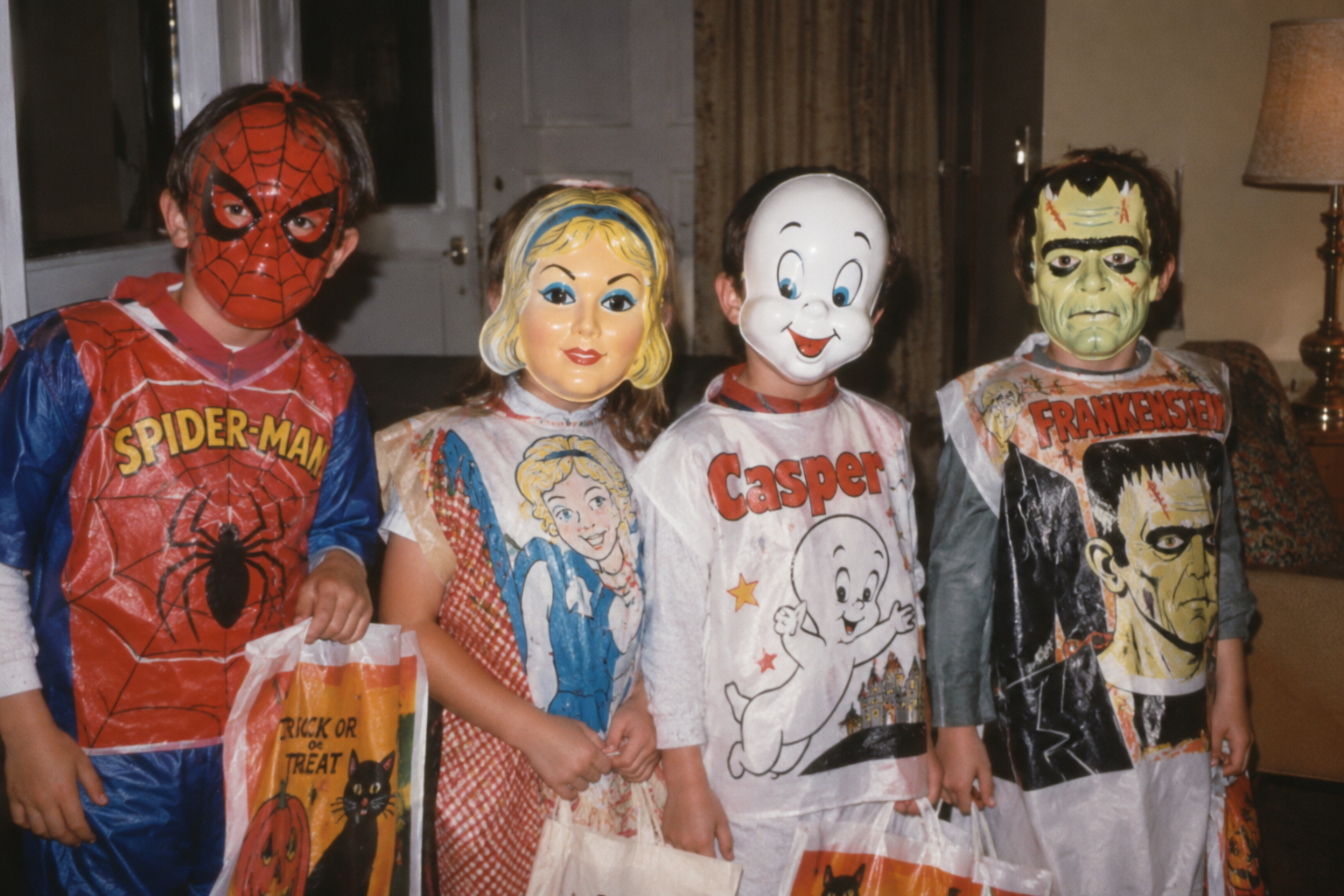 Four children dressed in vintage plastic Halloween costumes and masks representing Spider-Man, Cinderella, Casper the Friendly Ghost, and Frankenstein, holding trick-or-treat bags.