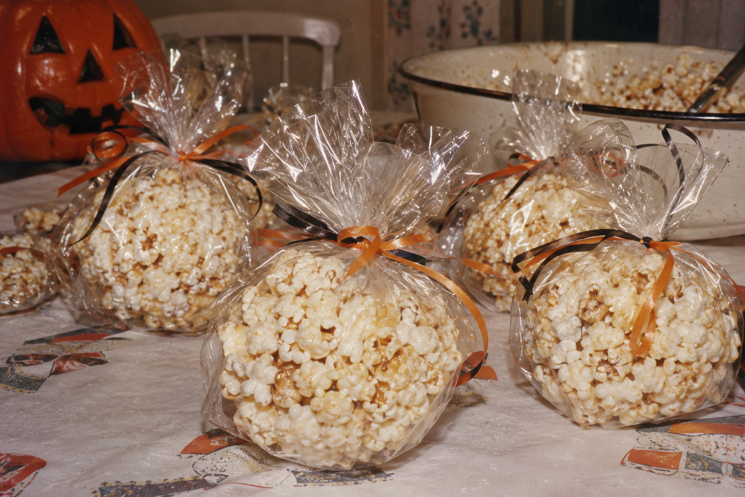Several popcorn balls wrapped in clear plastic bags with orange and black ribbons, sitting on a table next to a jack-o’-lantern.