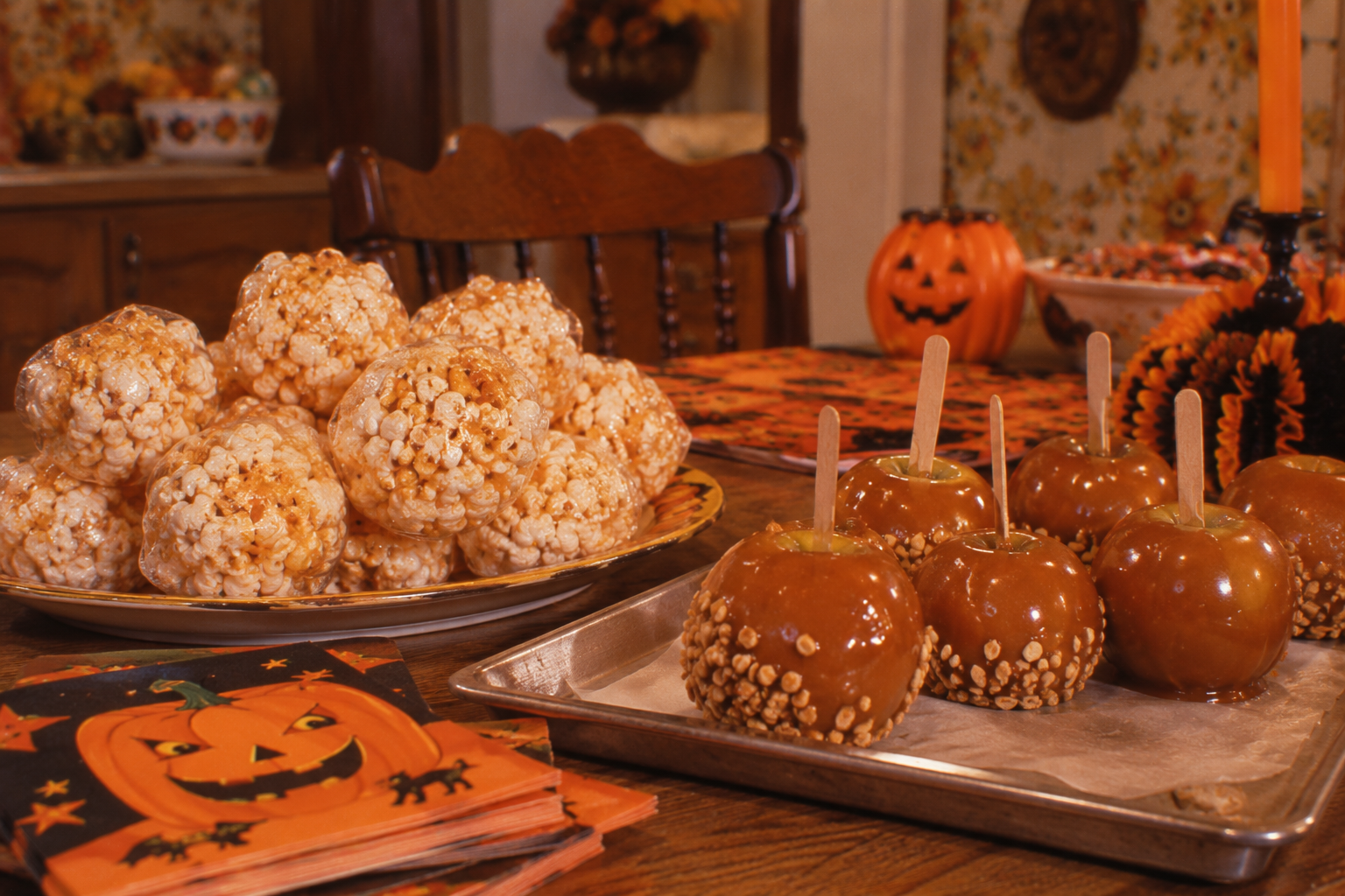 A platter of popcorn balls next to a tray of caramel apples topped with nuts, with Halloween-themed napkins in the foreground.