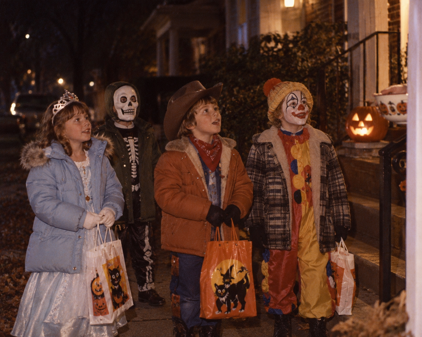 Four children in Halloween costumes—a princess, a skeleton, a cowboy, and a clown—standing on a porch with treat bags as they wait at a doorway with a jack-o’-lantern.