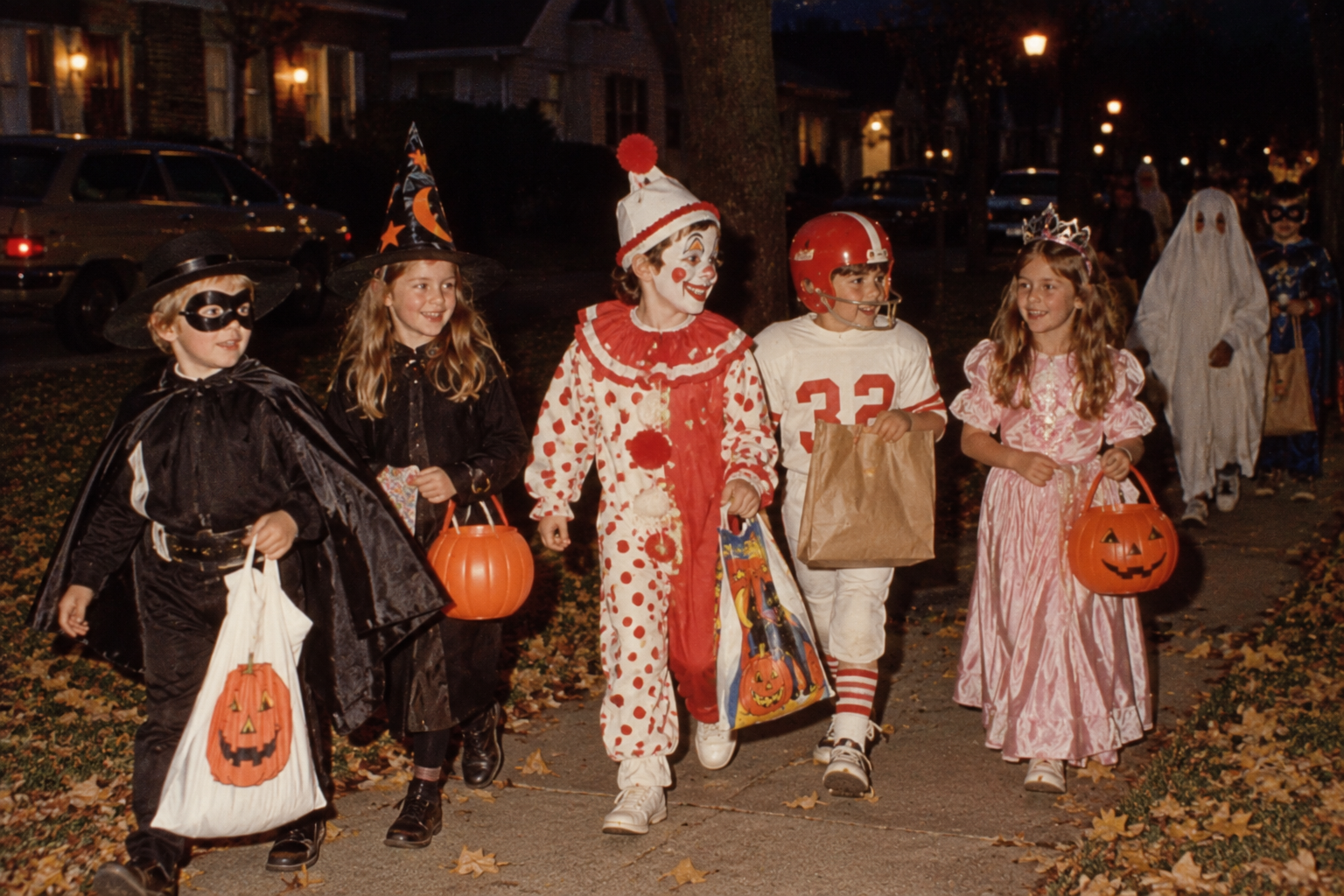 Five children in various costumes, including a Zorro-like character, a witch, a clown, a football player, and a princess, walk down a sidewalk at night while trick-or-treating.
