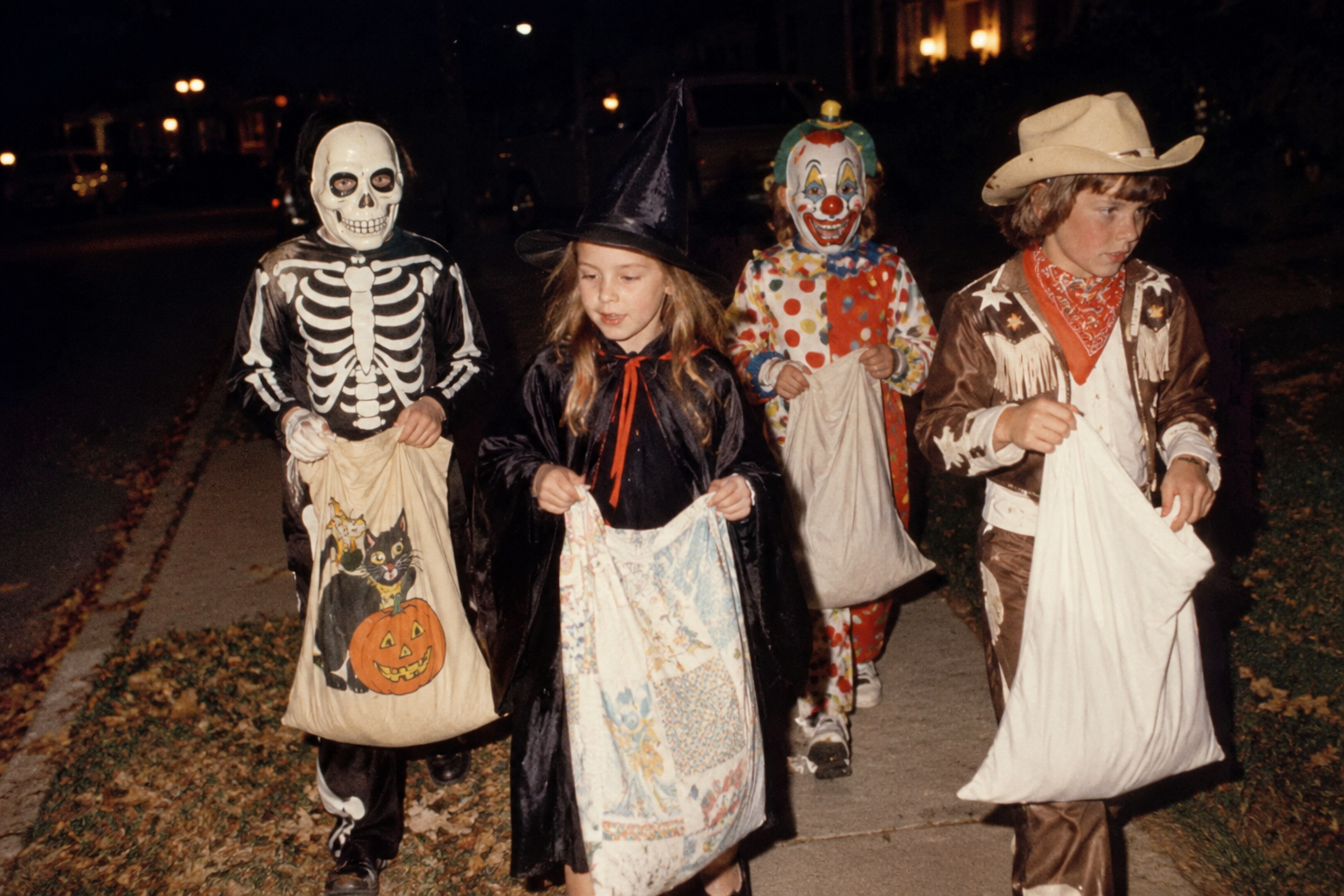 Four children in Halloween costumes—a skeleton, a witch, a clown, and a cowboy—walking down a sidewalk at night while carrying treat bags.