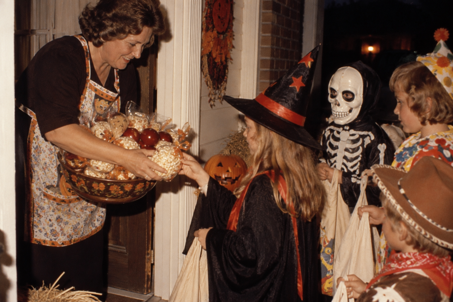 A woman in an apron hands treats from a bowl to children in Halloween costumes standing on a porch.