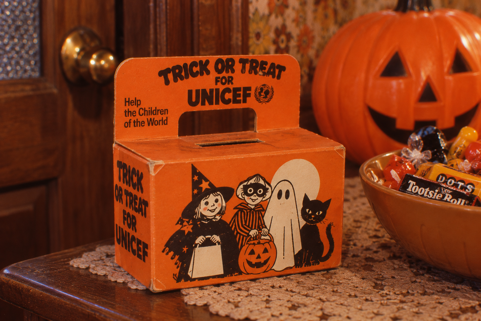Orange cardboard UNICEF Trick-or-Treat donation box illustrated with a witch, a masked child, a ghost, and a black cat, sitting on a table next to a bowl of candy and a jack-o’-lantern.