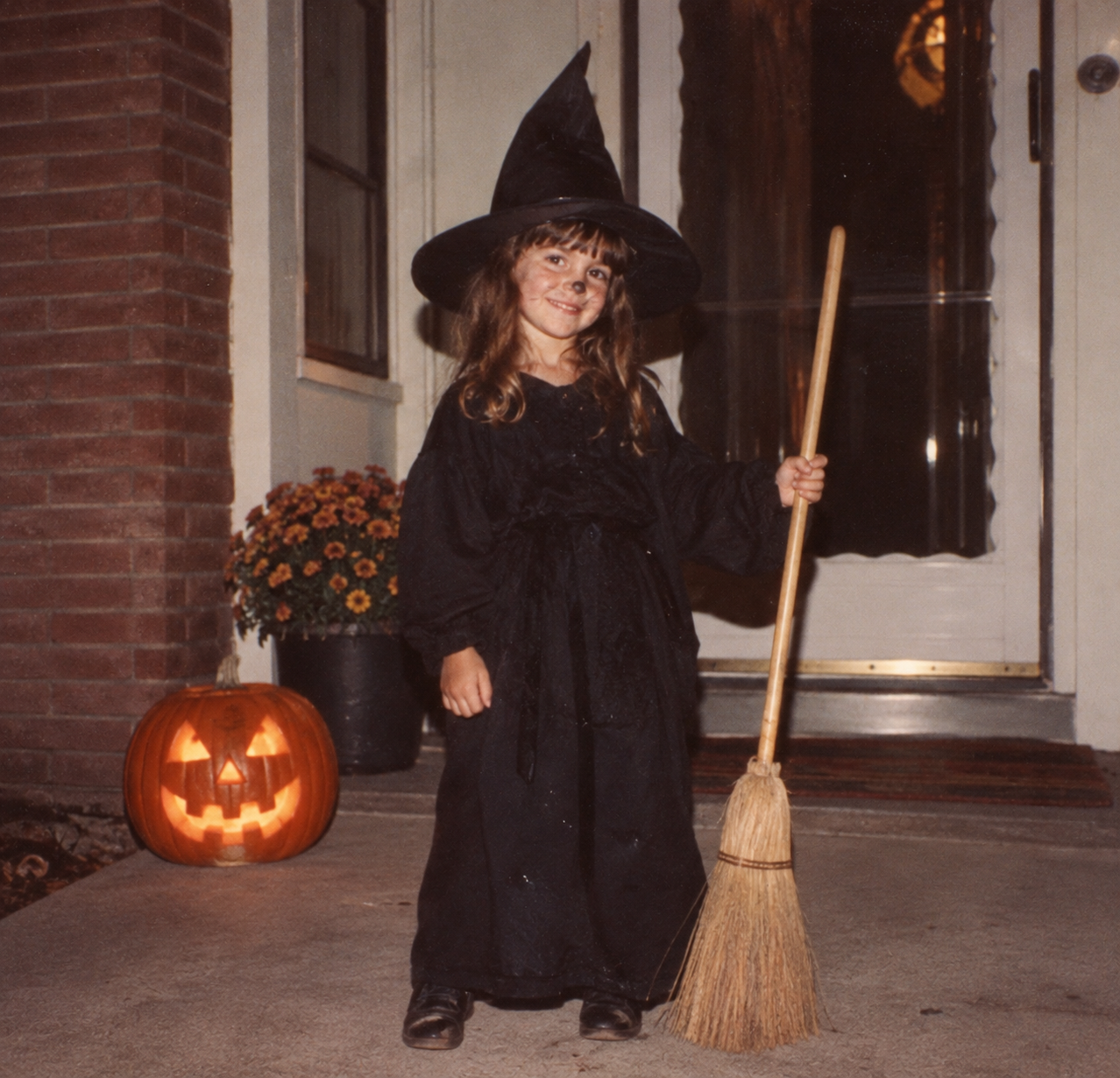 A young girl in a black witch costume and pointed hat stands on a porch next to a glowing jack-o’-lantern while holding a broom.