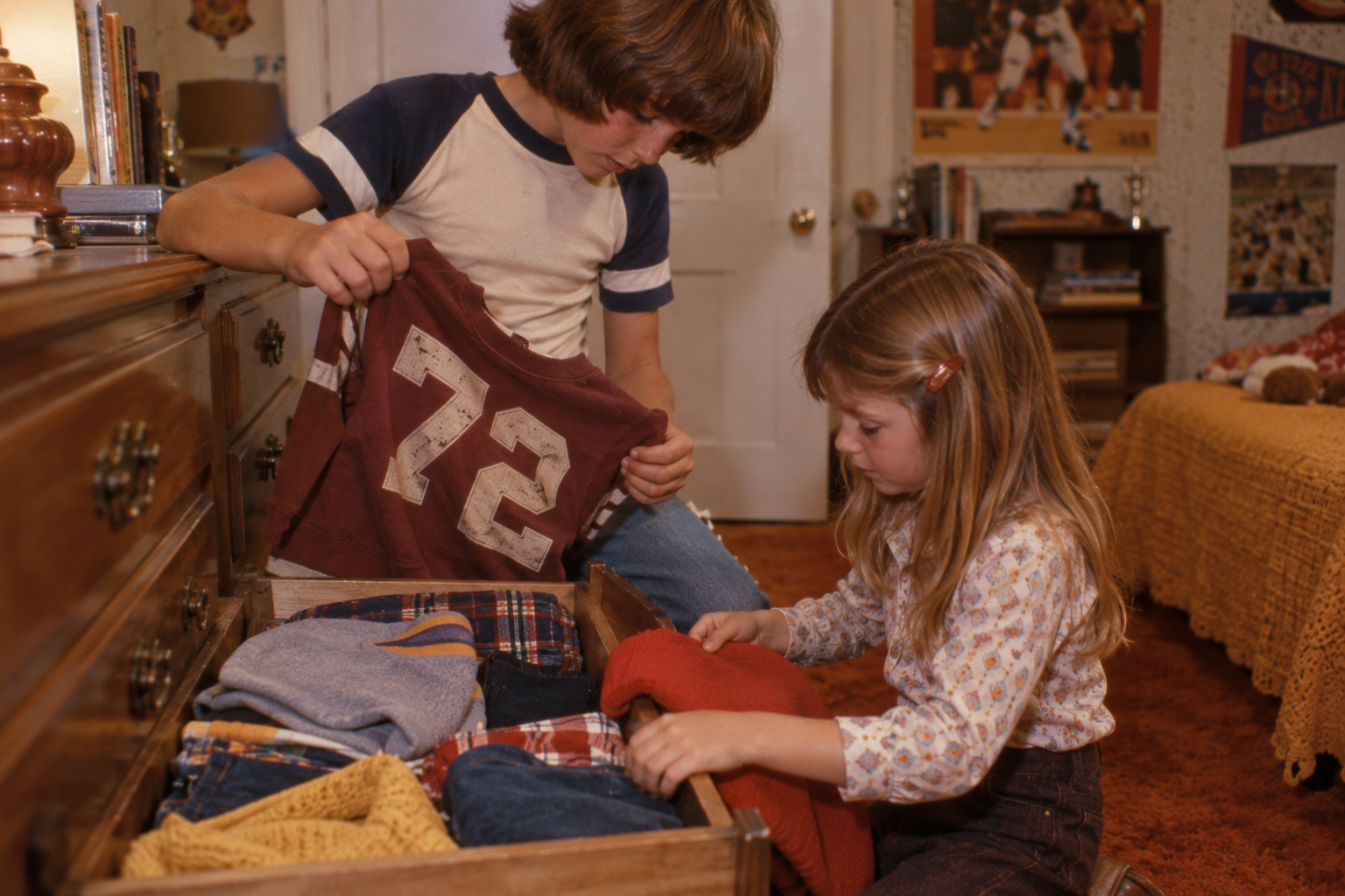 A young boy and girl look through a wooden dresser drawer filled with folded clothes.