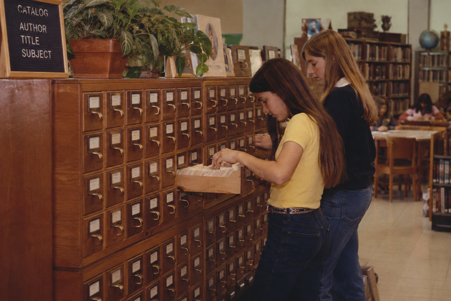 Two teenage girls searching through wooden card catalog drawers in a library.