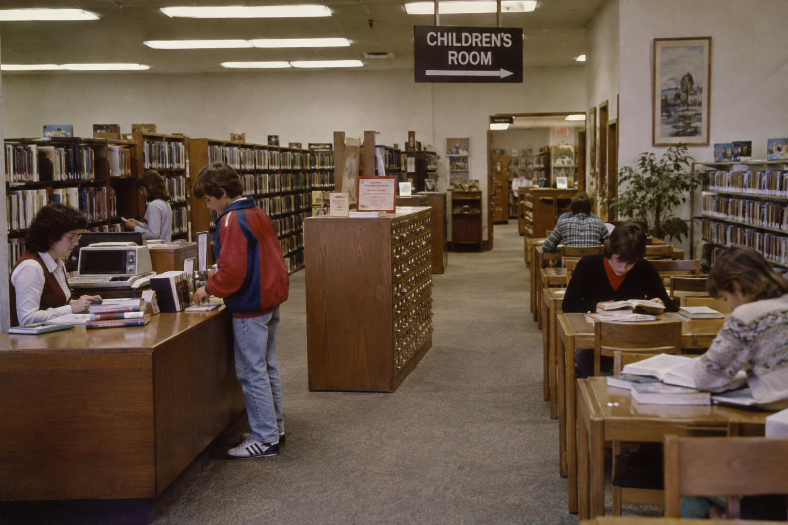 Patrons browse and study in a public library, with a sign pointing toward the children’s room. Places ’80s kids hung out.