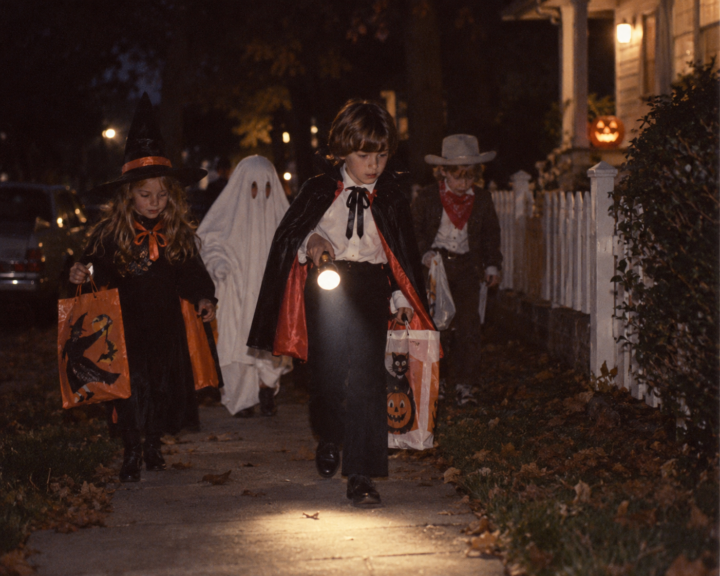 Four children in Halloween costumes walk along a sidewalk at night, with one child holding a flashlight. Halloween in the 1970s.