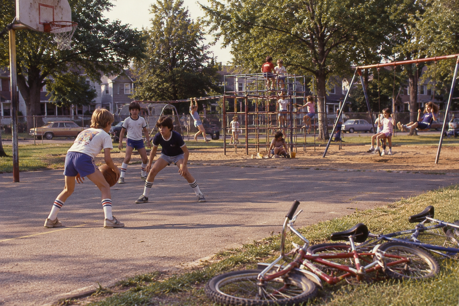Two boys play basketball on an outdoor court, while other children play on a nearby jungle gym and swings, with bicycles lying on the grass in the foreground.