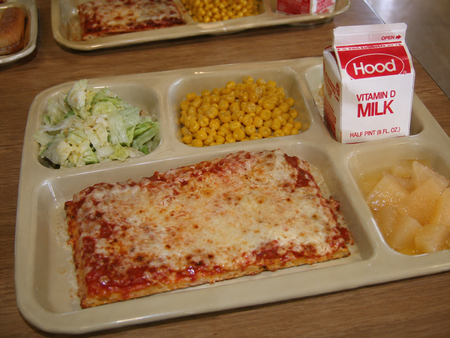 School lunch tray with a rectangular slice of cheese pizza, a serving of lettuce salad, corn, diced fruit, and a carton of Hood Vitamin D milk. 1980s school cafeteria.