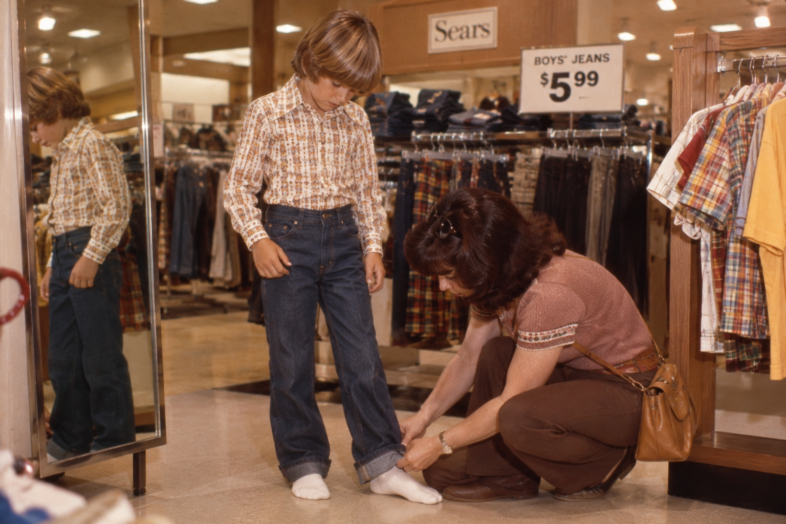 A woman kneels to cuff the hem of a boy’s jeans in a department store.