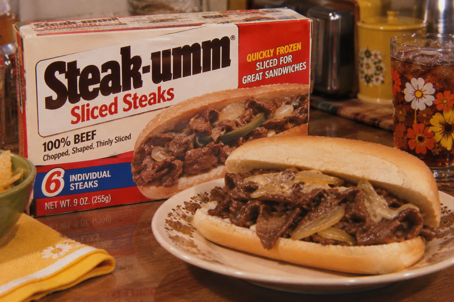 A box of Steak-umm Sliced Steaks is next to a sandwich filled with cooked beef and onions on a plate. There is a glass of soda with ice and a floral design. The setting appears to be a kitchen or dining table.