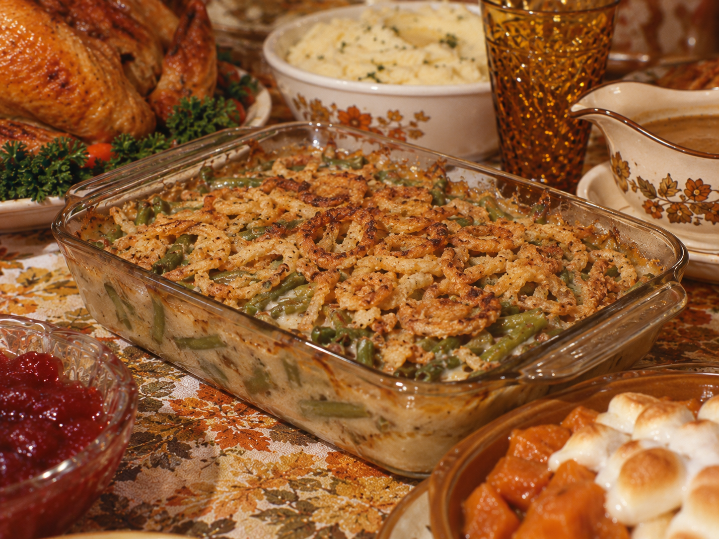 A glass baking dish of green bean casserole topped with fried onions, surrounded by dishes of roast turkey, mashed potatoes, gravy, cranberry sauce, and sweet potatoes with marshmallows on a floral tablecloth.