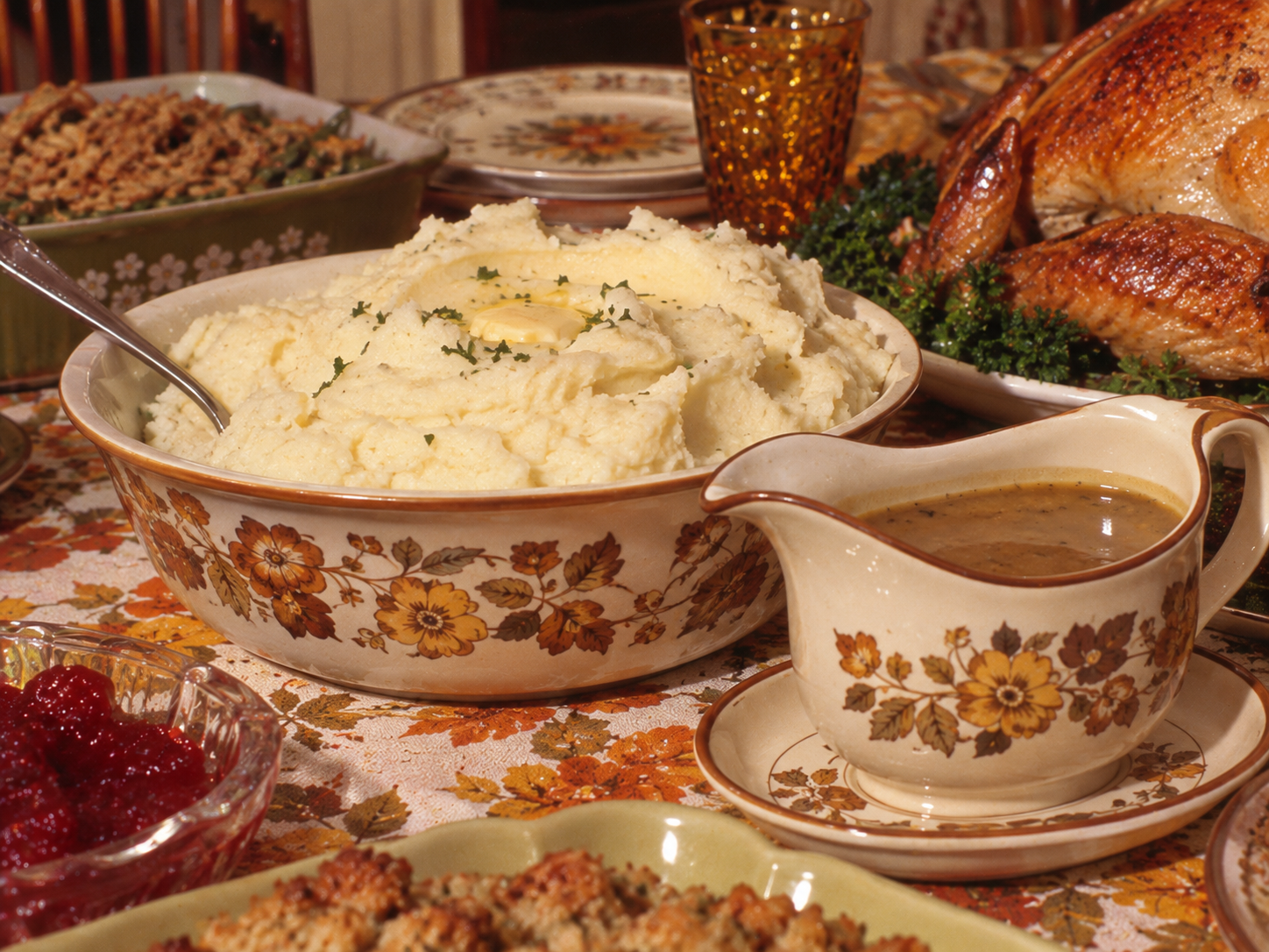 A Thanksgiving dinner table featuring a large bowl of mashed potatoes with butter, a roasted turkey, gravy in a floral boat, cranberry sauce, and stuffing.