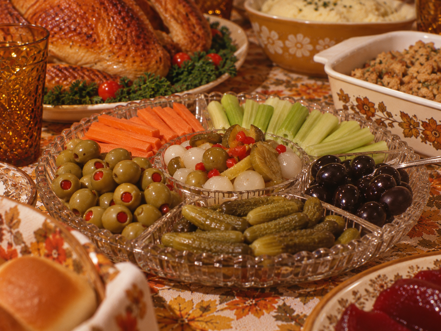 A crystal relish tray containing carrot sticks, celery sticks, green pimento-stuffed olives, black olives, pickles, and pickled onions, set on a table with other Thanksgiving dishes.