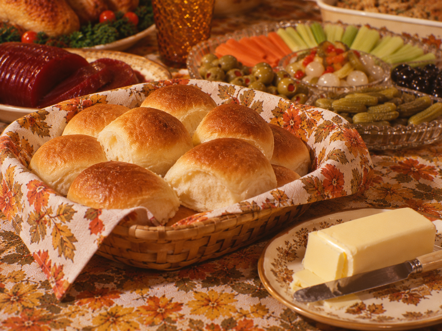 A wicker basket filled with golden-brown dinner rolls, next to a plate with a stick of butter and a knife, set on a table with other holiday dishes.