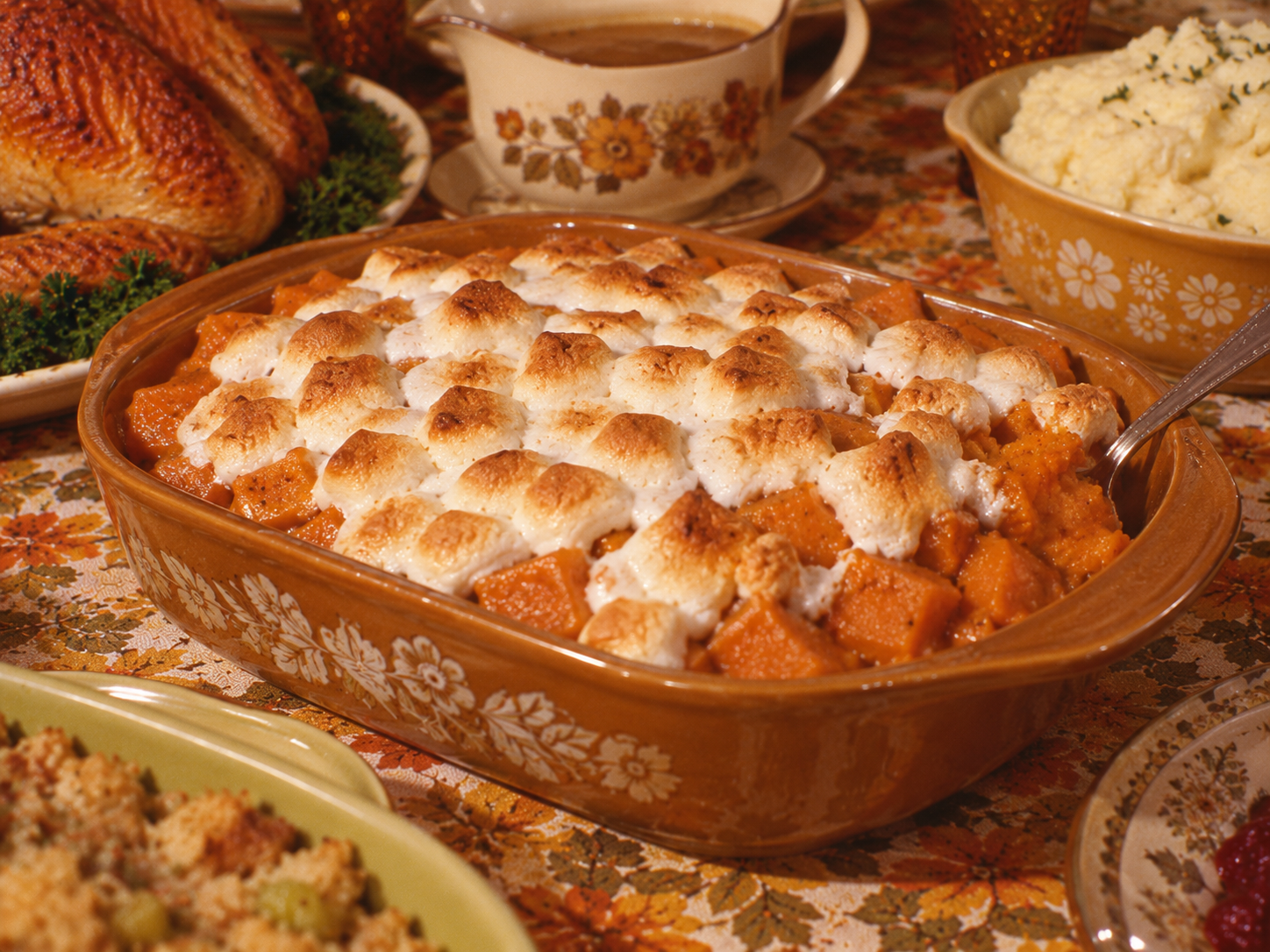 Casserole dish filled with cubed sweet potatoes topped with toasted marshmallows, surrounded by Thanksgiving side dishes.